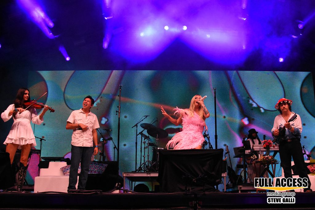 Ensemble performing with singers and a violinist under purple festival lights