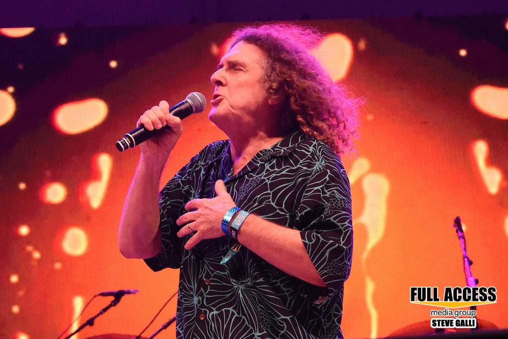 Curly-haired vocalist singing with one hand on their chest