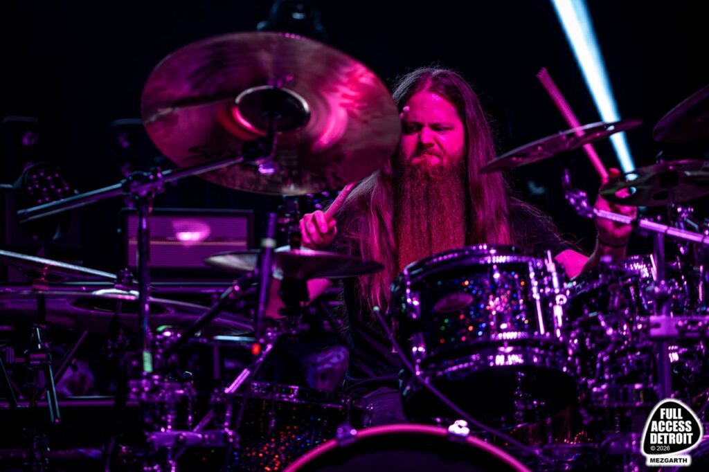 Long-haired drummer playing a drum kit under purple stage lighting