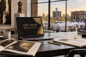 Laptop displaying the Art Show Magazine name beside editorial proofs, with an outdoor art fair and Detroit skyline beyond a gallery window.